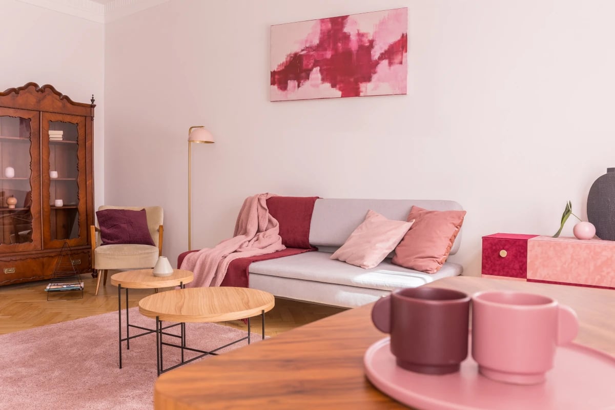 Pomieszczenie z kanapą oraz fotelem. Narzuty, koce oraz poduszki w kolorze różowym. Pod ścianą stoi kredens. Na środku stoją stoliki kawowe z drewnianym blatem.