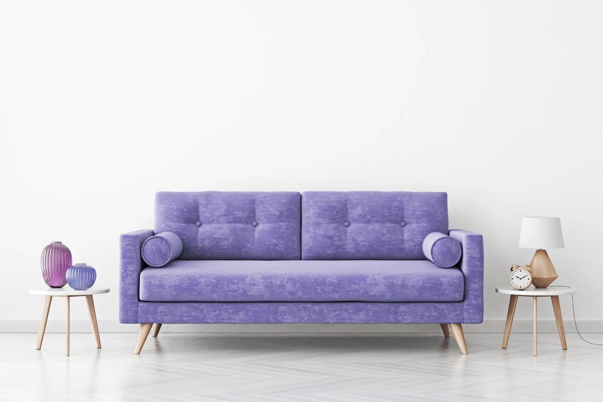 Fioletowa kanapa na tle białej ściany. Z boku kanapy stoją dwa stoliki z dekoracjami w odcieniach fioletu.