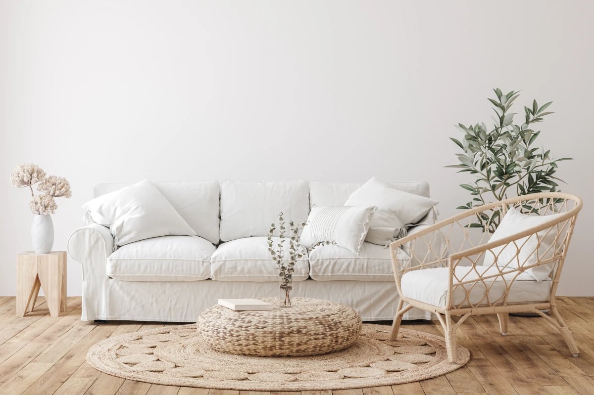 Ażurowy fotel stojący przy jasne kanapie na tle białej ściany. Na podłodze dywan okrągły. Obok kanapy w stoliku z wazonem i kwiatami.