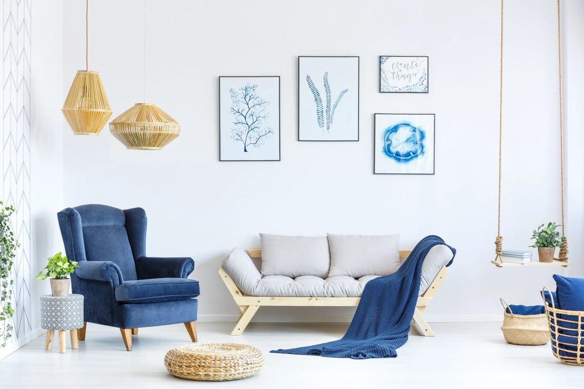 Drewniana konstrukcja sofy z szarymi poduszkami na tle białej ściany. Fotel z tapicerką w kolorze niebieskim. Na ścianie obrazy z niebieskimi akcentami. 