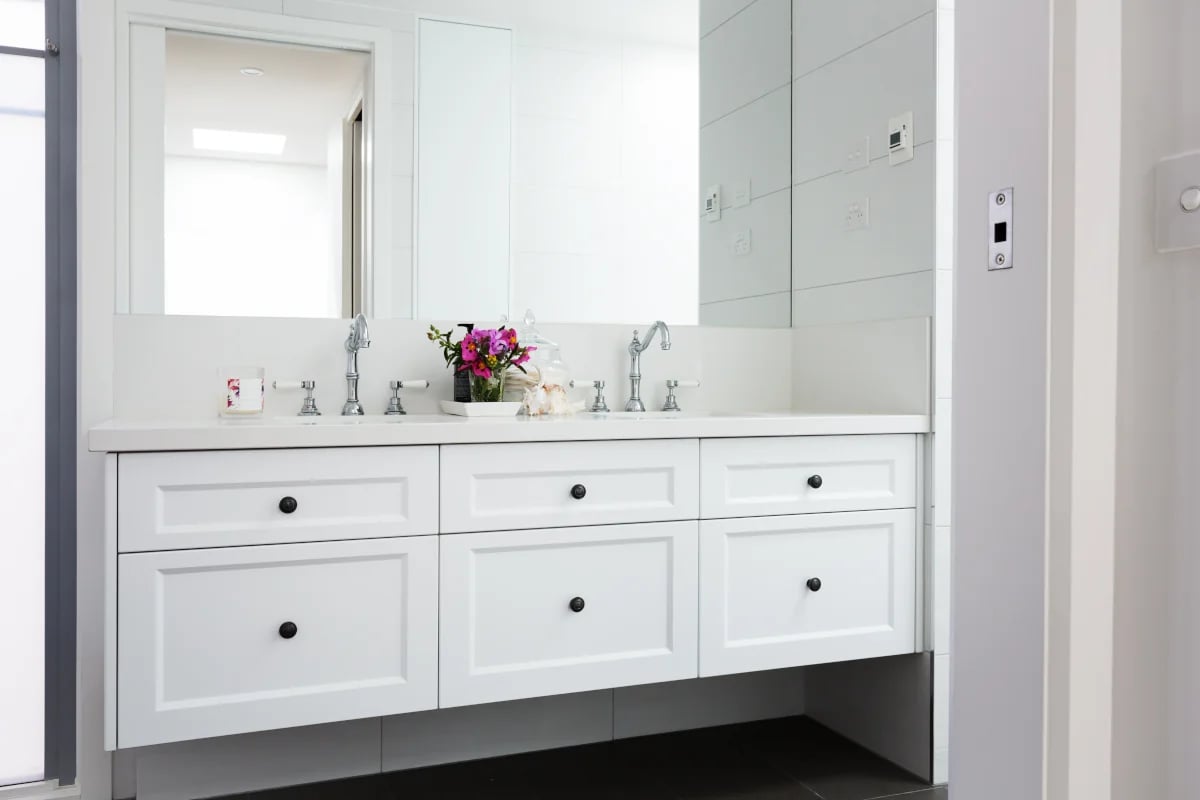 Białe meble łazienkowe z czarnymi gałkami. Dwie umywalki wpuszczone w blat oraz lustro nad nimi.