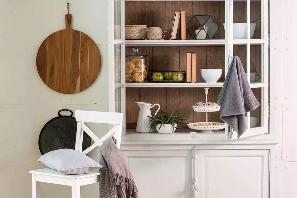 Starodawnym kredens z akcesoriami kuchennymi oraz dekoracjami. Biała krzesło obok kredensu z poduszką oraz narzutą.