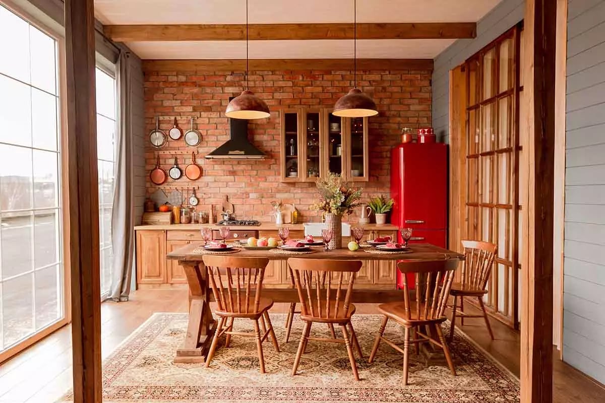 Kuchnia w połączeniu z jadalnią. Ściana z meblami z czerwoną cegiełką. Stół, krzesła oraz meble kuchenne drewniane. Czerwona lodówka stoi w kącie. Duże okno w pomieszczeniu.
