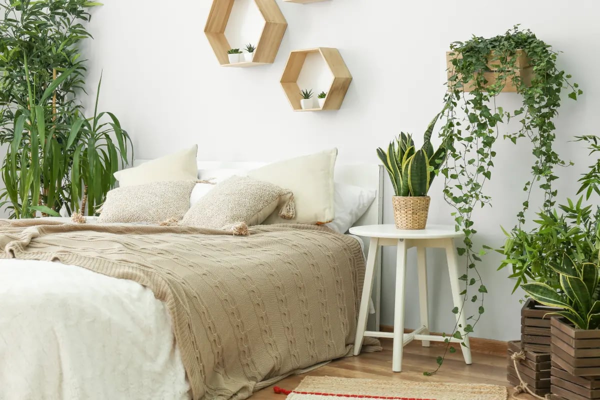 Łóżko z narzutą oraz poduszkami w kolorze beżu. Na ścianie drewniane półki. Wokół dużo kwiatów.