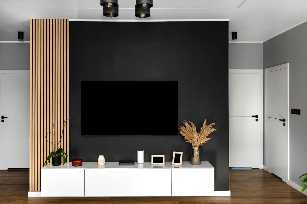 Ścianka z telewizorem w mieszkaniu. Z lewej strony telewizora drewniane lamele. Pod telewizorem biała szafka.