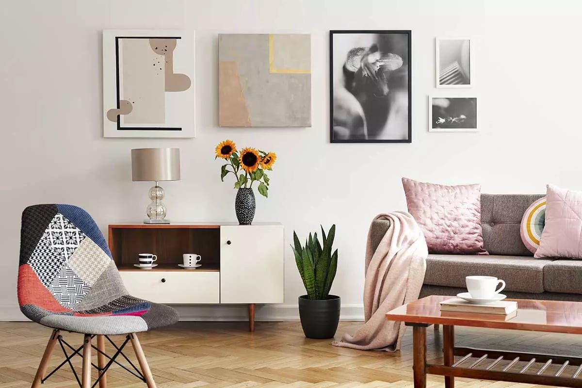 Kanapa z poduszką na tle białej ściany z obrazami oraz zdjęciami. Pod ściana szafka, a na środku krzesło z kolorowym obiciem.