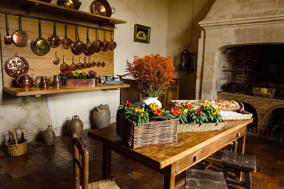 Kuchnia w stylu rustykalnym. Stół drewniany z koszykami wiklinowymi z owocami oraz pieczywem. Z boku półka oraz wieszaki na miedziane garnki oraz czajniki. Z prawej strony piec.
