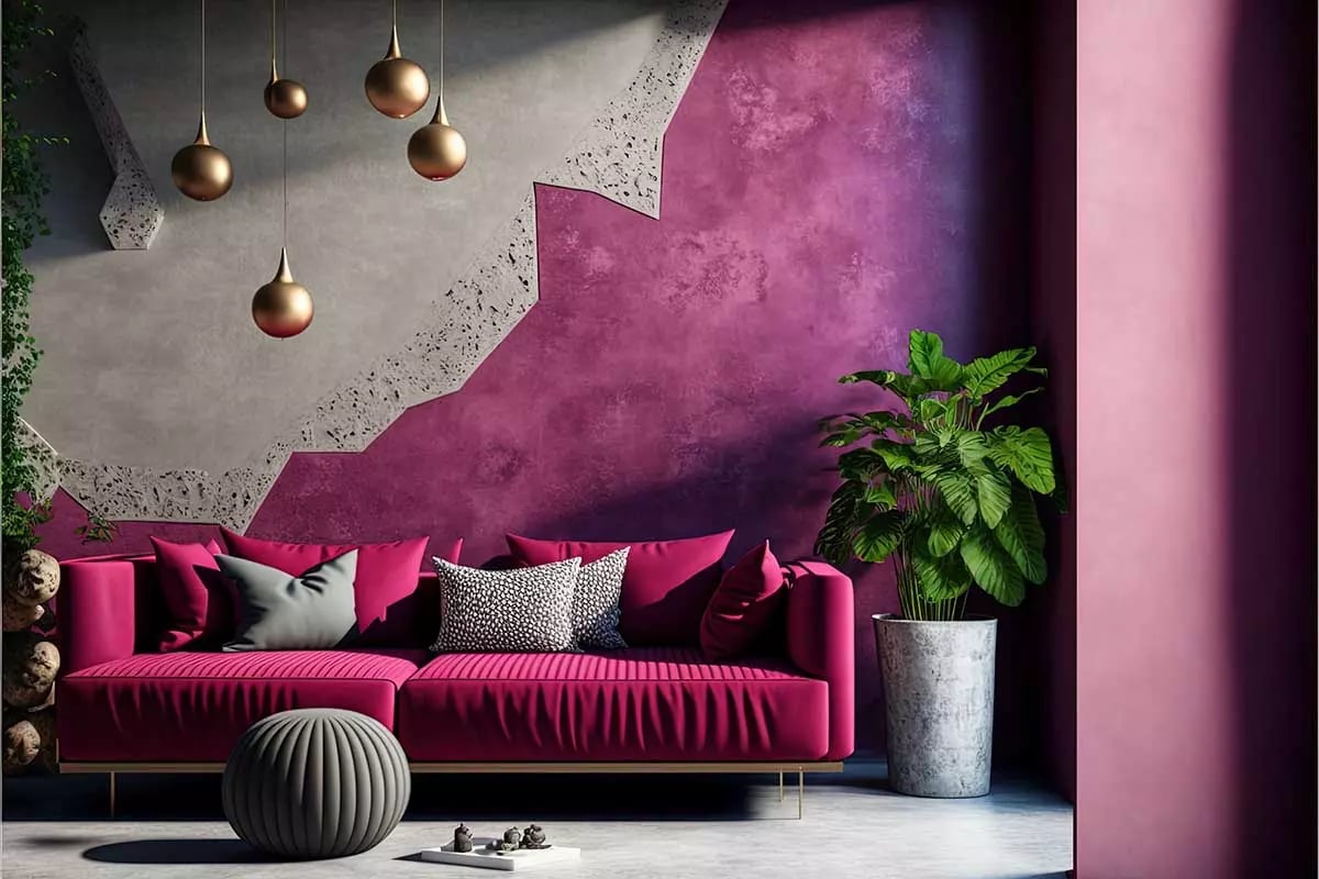 Kanapa w kolorach różu z poduszkami, na tle fioletowej ściany połączonej z akcentami betonowymi. Obok kanapy donica z kwiatem.