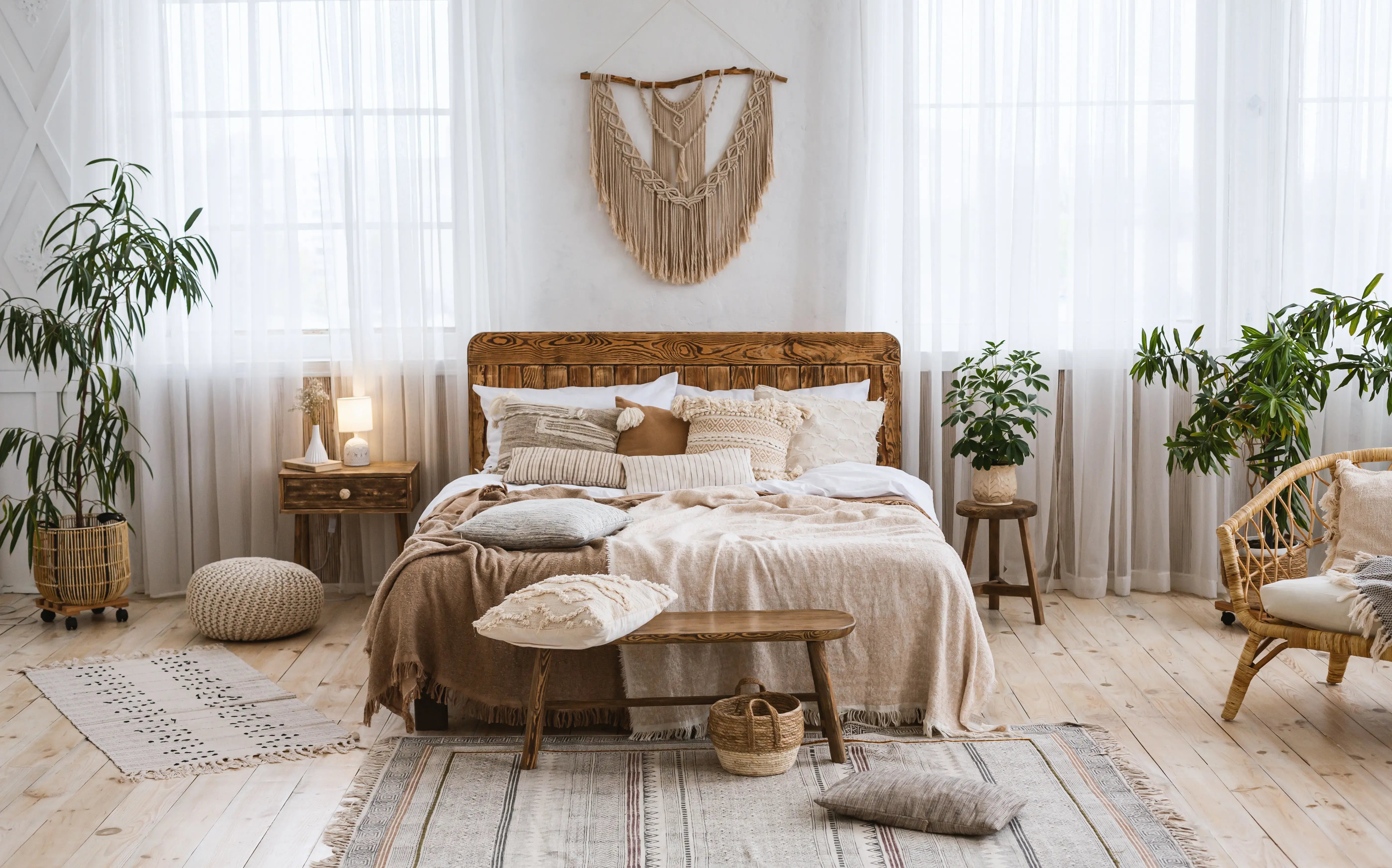 Sypialnia w stylu scandi boho z kolorze białym  z dużym drewnianym łóżkiem w centrum, markamą nad nim, obok łóżka szafka nocna, na której stoi lampka, a po drugiej stronie krzesło, na którym stoi doniczka z kwiatkiem. 