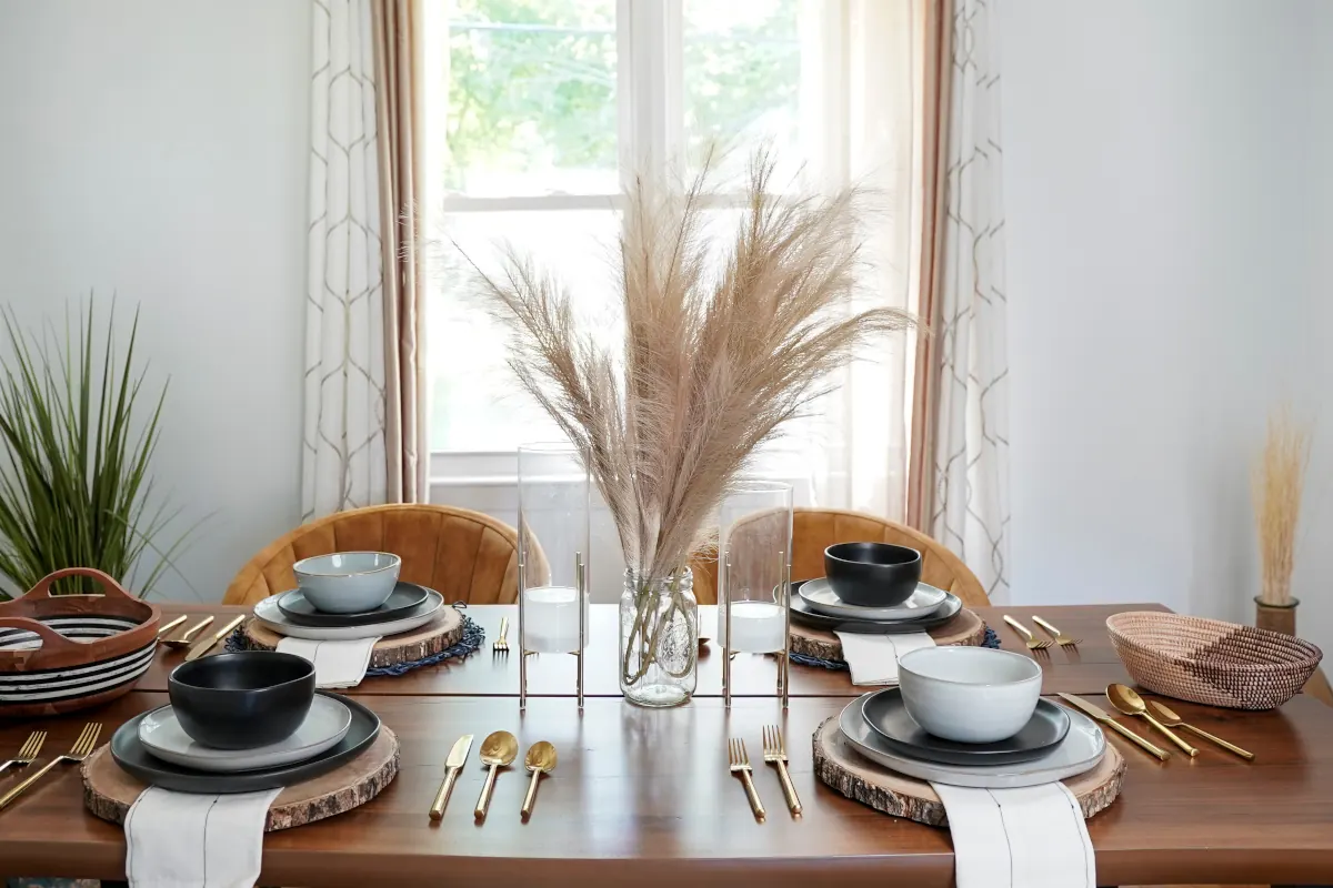 Zastawiony stół talerzami na drewnianym podkładkach. Na stole znajdują się dekoracje w stylu boho oraz sztućce w stylu glamour.