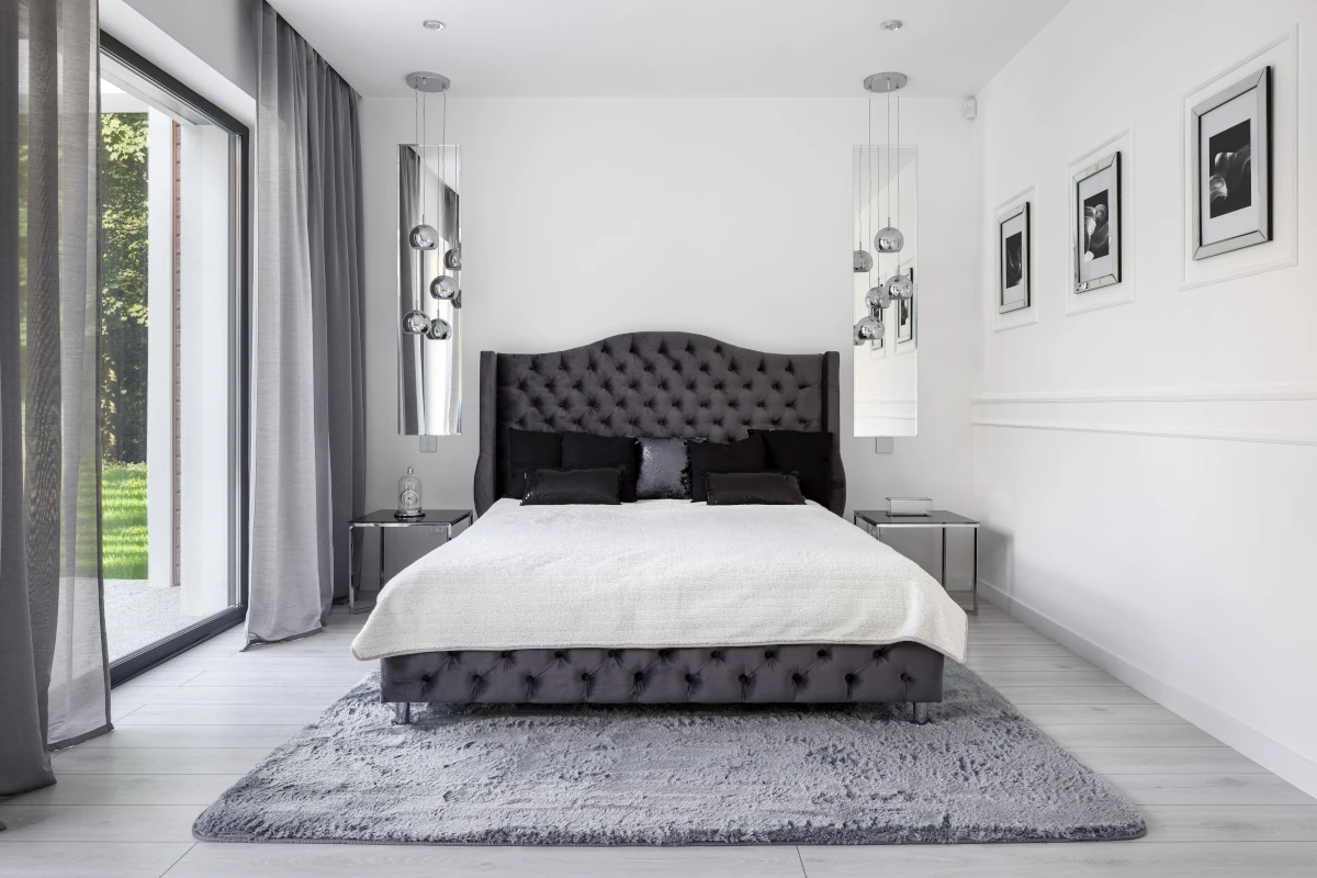 Sypialnia w kolorach szarości oraz bieli. Łóżko ustawione na środku pomieszczenia. Dodatki nadają pomieszczeniu styl glamour.
