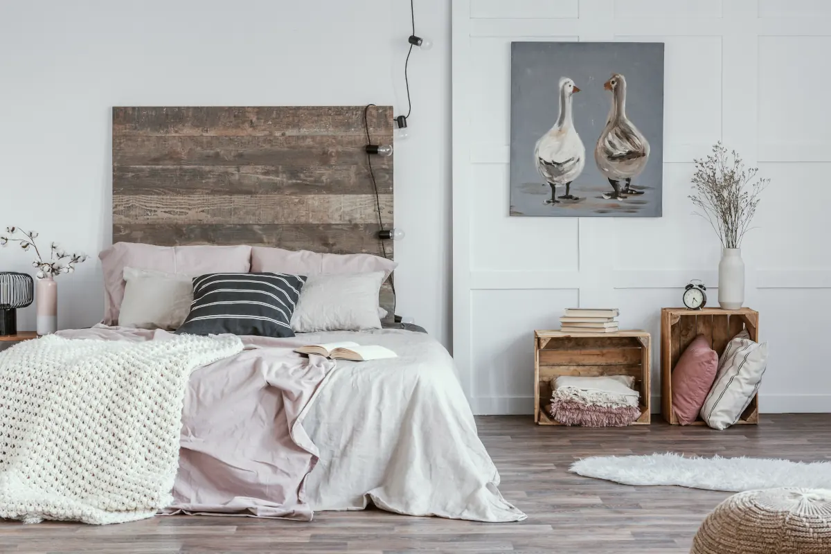 Łóżko z narzutą i poduszkami. Obok stoją drewniane skrzynki. Obraz wisi na białej ścianie.