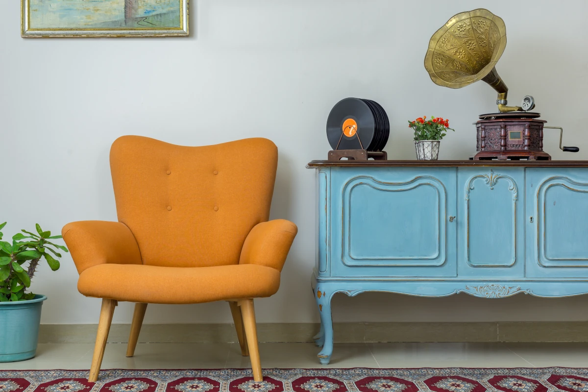Błękitna szafka ze żłobieniami. Obok stoi fotel z pomarańczowym obiciem. Na szafce stoi gramofon oraz płyty winylowe.