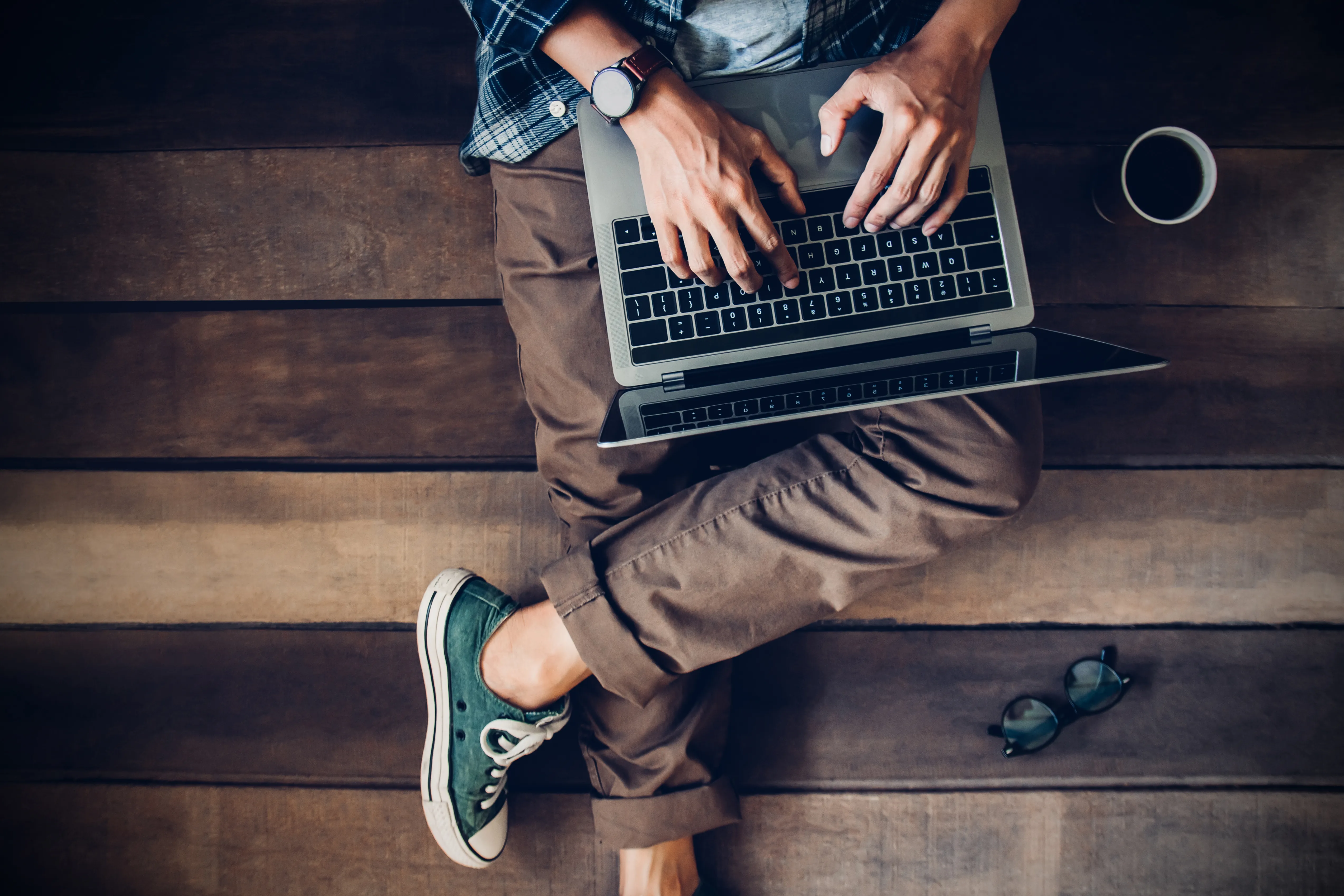 Mężczyzna siedzi na drewnianych schodach z laptopem na kolanach, jego ręce są na klawiaturze laptopa i coś wyszukują.