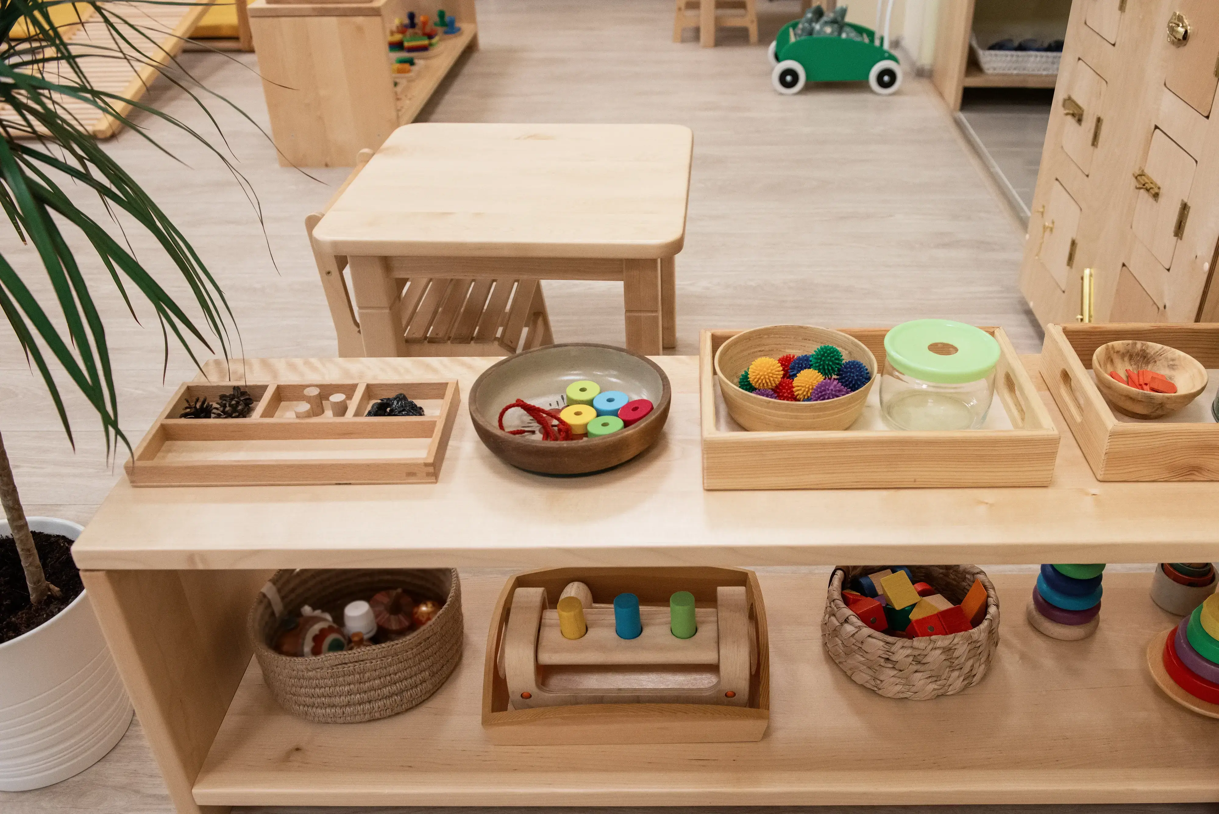 Pokój w przedszkolu zaaranżowany w stylu Montessori, gdzie widzimy dużo drewnianych mebli, a na małym regale leżą zabawki ułożone w różnych miskach.