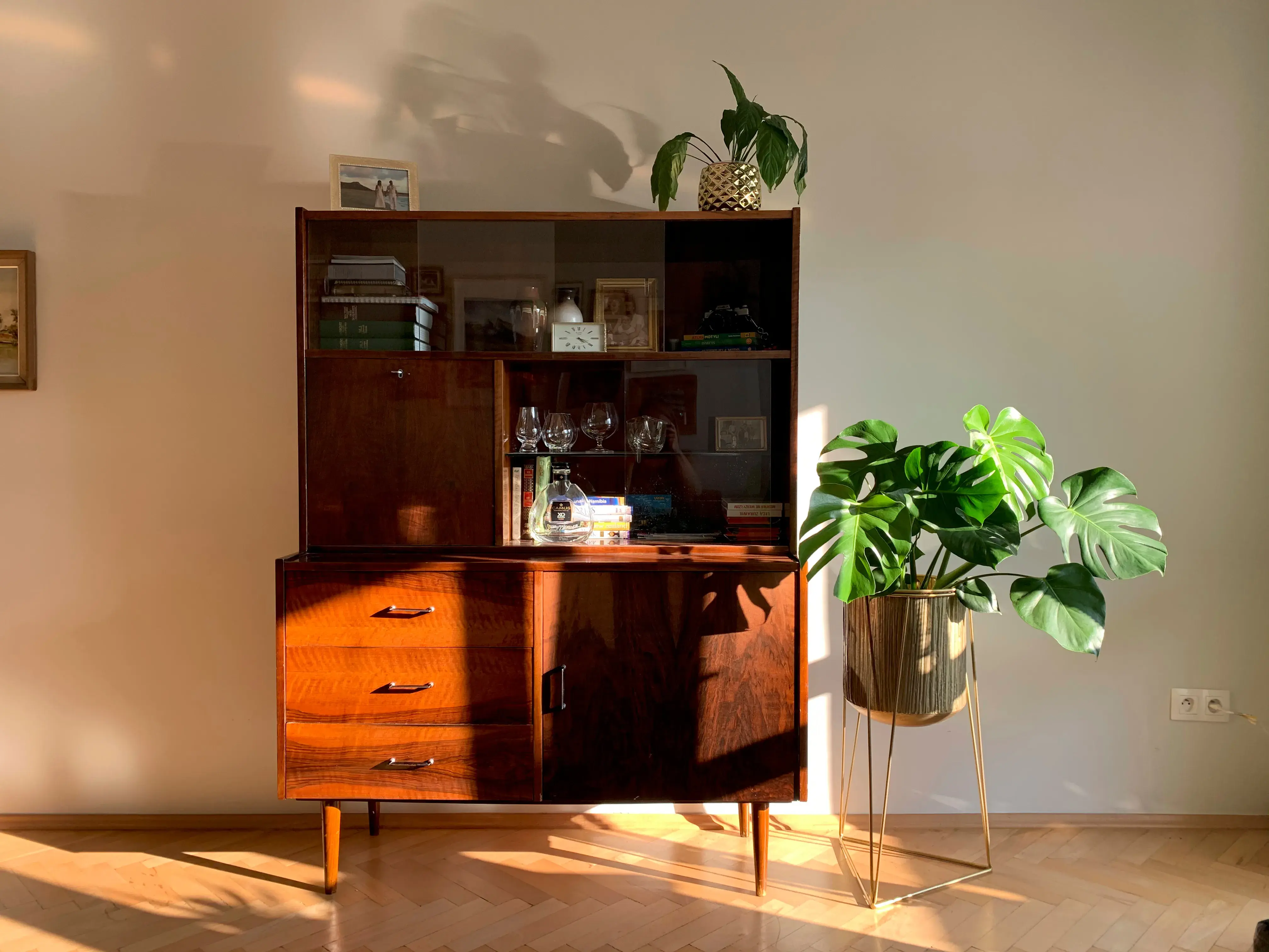 Salon w stylu PRL z białymi ścianami, drewnianą posadzką i dużą brązową szafką przypominająca czasu PRL-u.