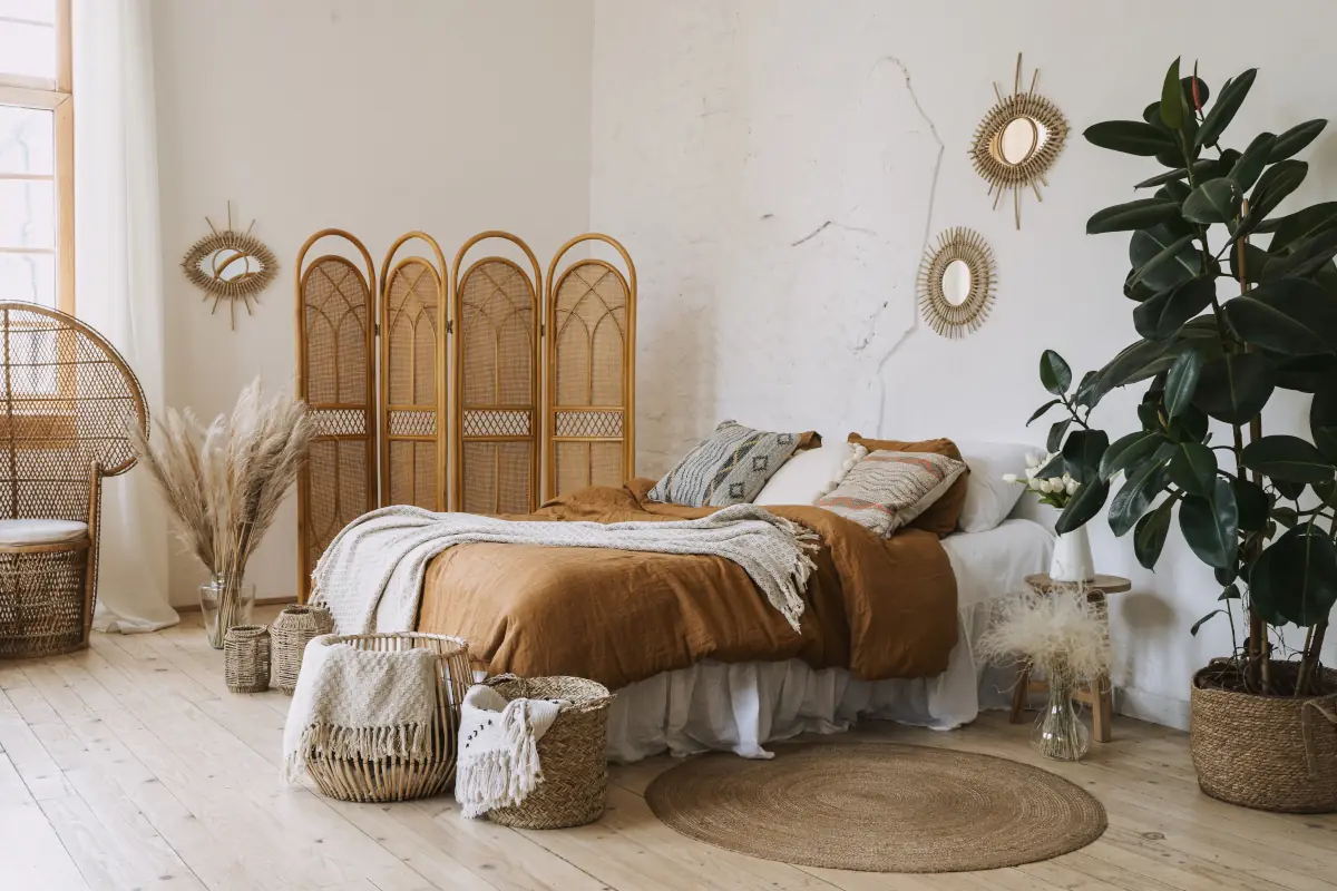 Łóżko w sypialni z narzutami w kolorze ziemi. Przy łóżku znajdują się kosze wiklinowe oraz rośliny w osłonach wiklinowych.
